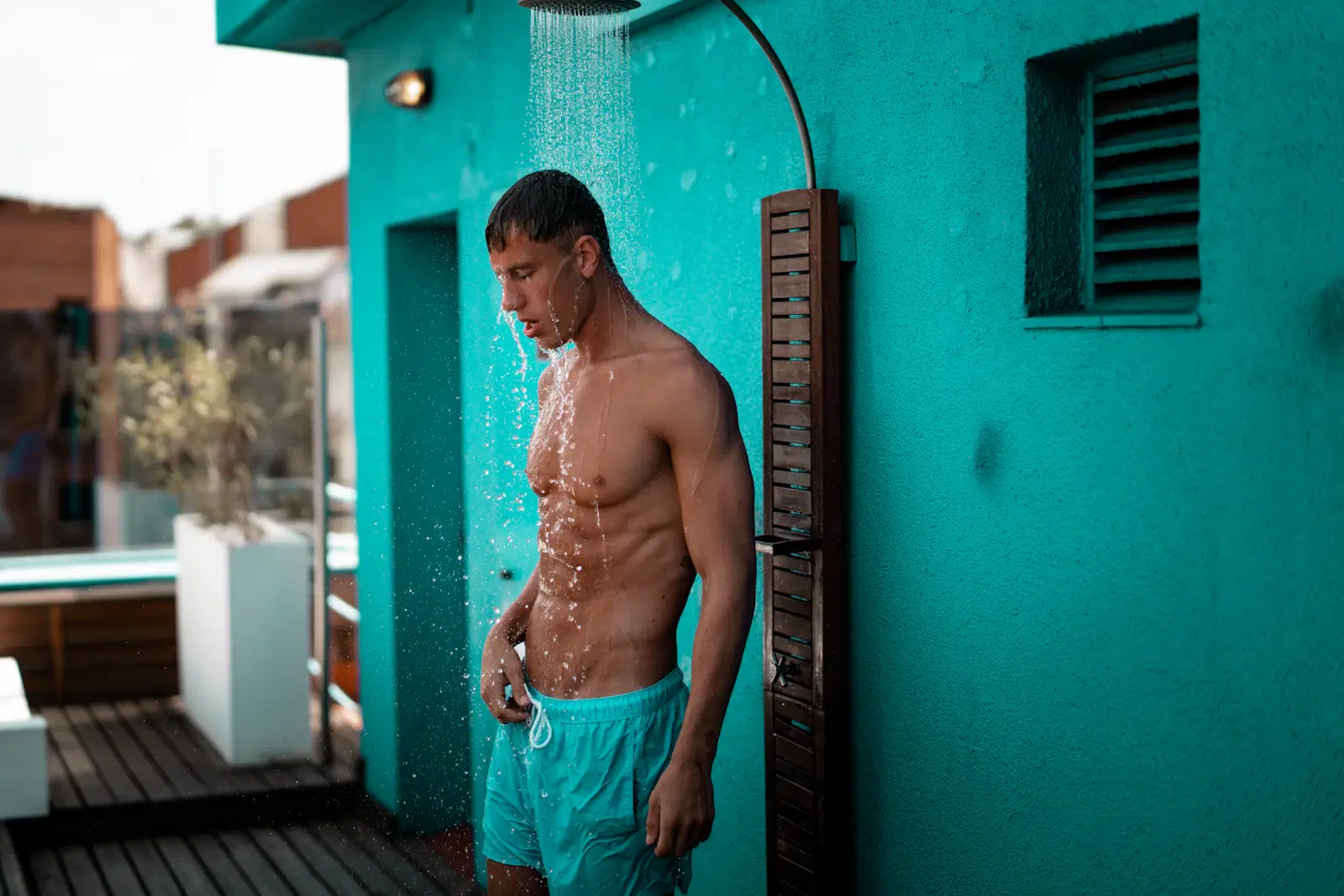 A man rinsing off under an outdoor shower against a teal wall