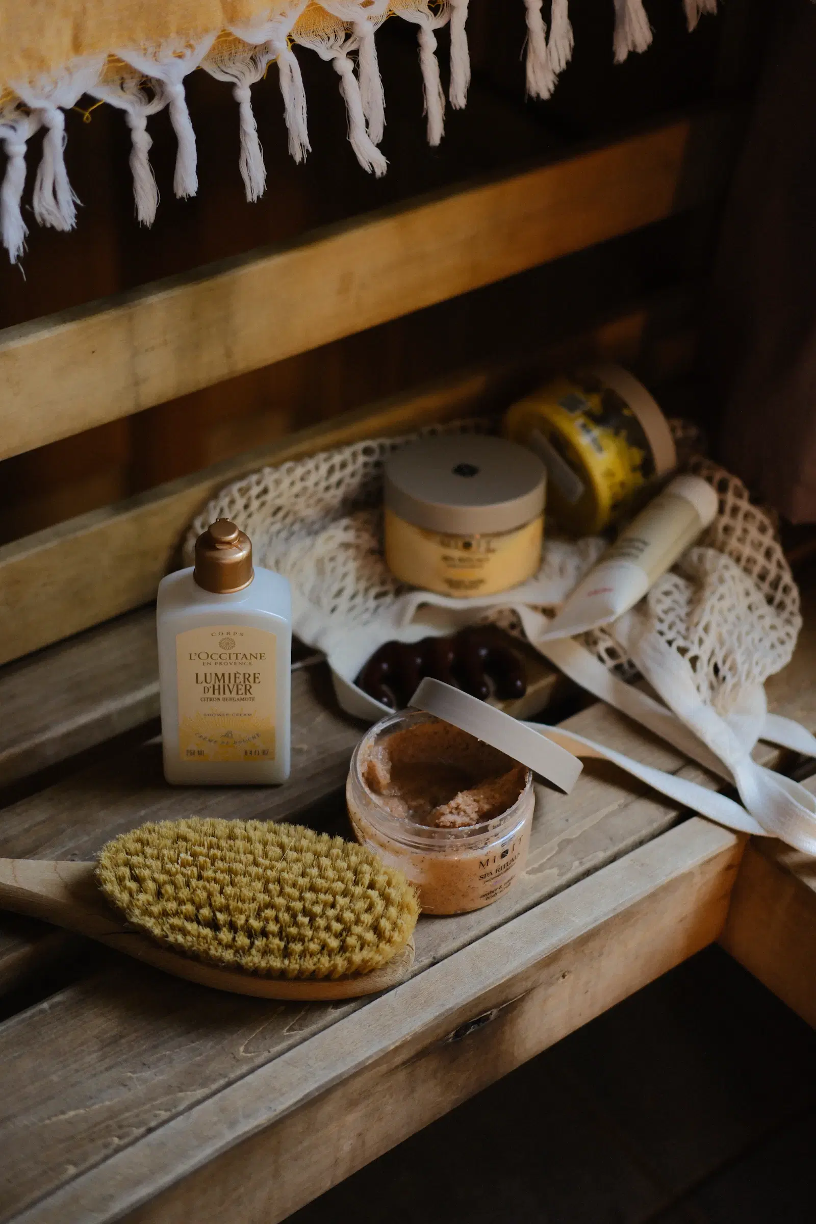 A wooden dry body brush next to a body scrub jar and shower cream on a wooden bench