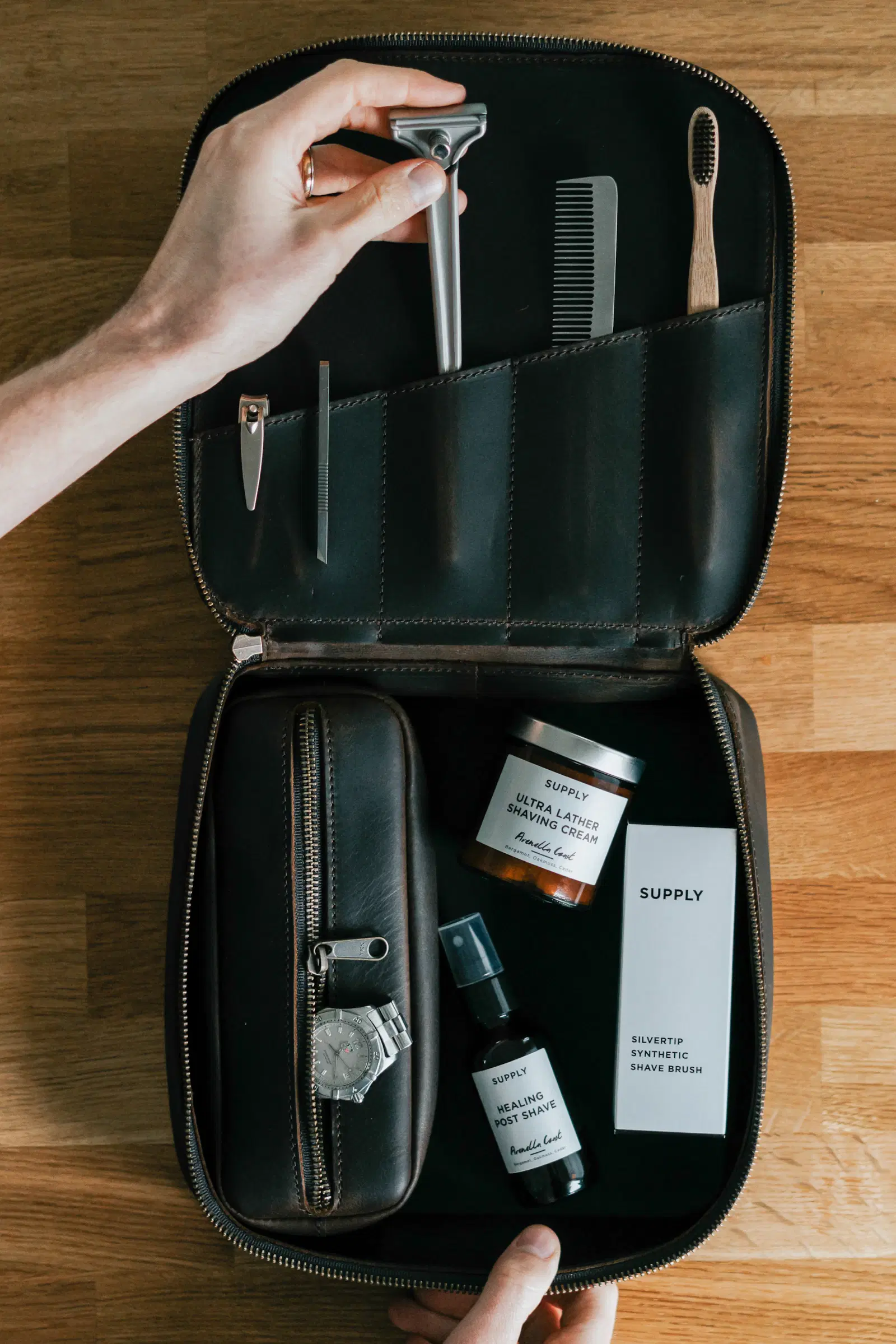 A hand taking grooming tools from a leather travel kit with a razor, comb, and skincare