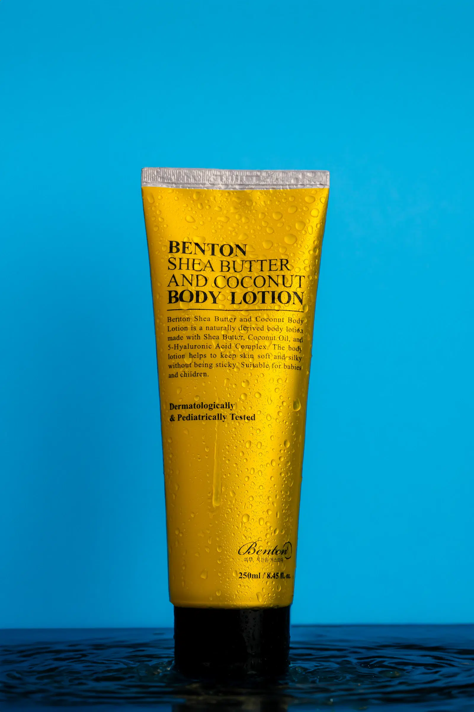 A tube of shea butter body lotion standing in shallow water against a blue backdrop