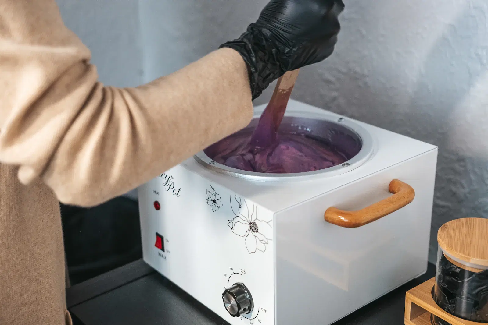 A gloved hand stirring purple hard wax in a wax warmer