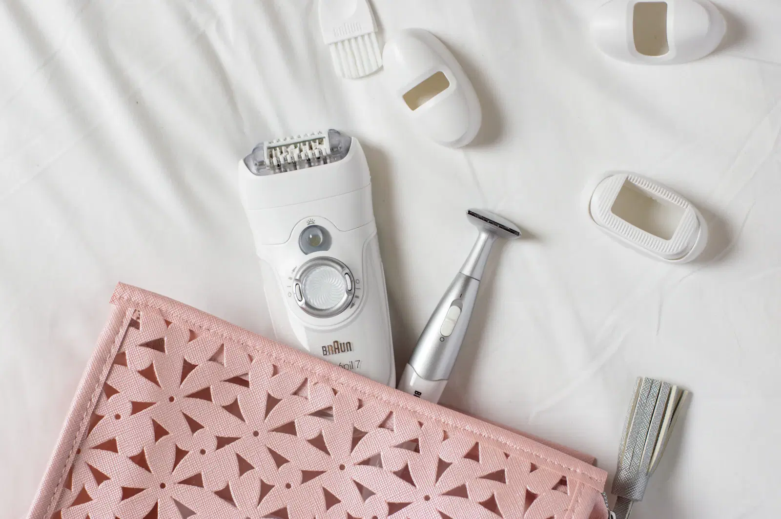 A white Braun Silk-épil 7 epilator with its caps, cleaning brush, and a pink travel pouch on a white sheet
