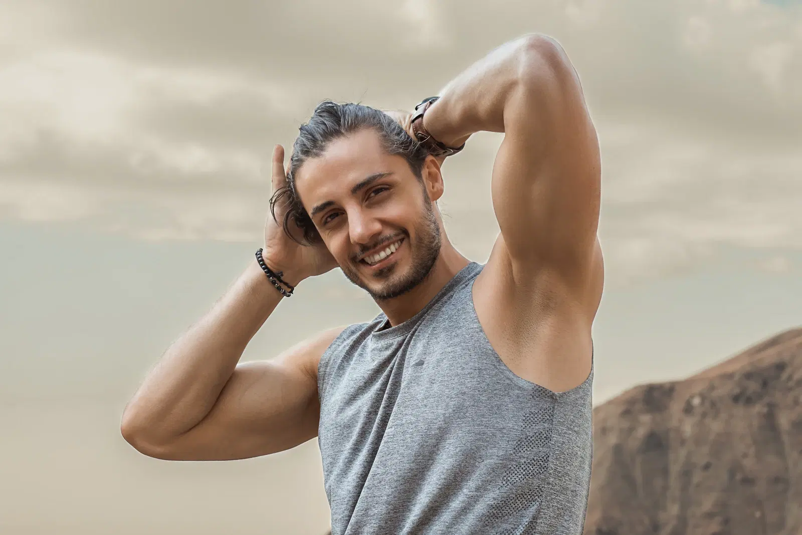 A smiling man in a gray tank top with his arms raised behind his head