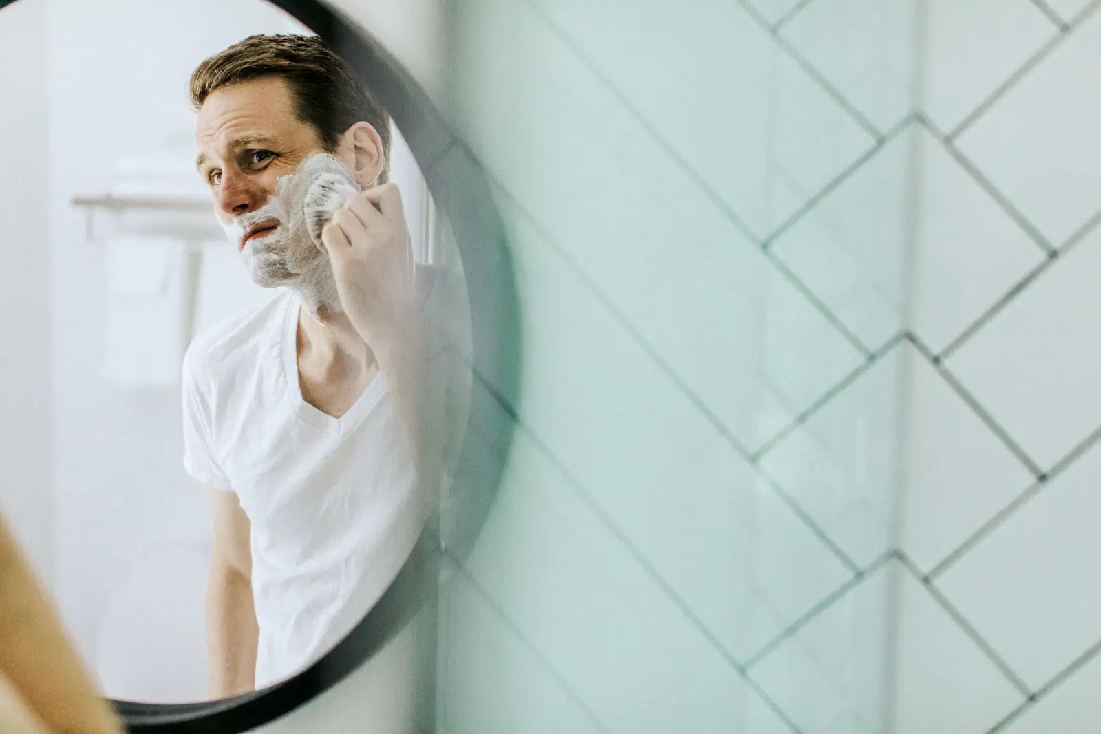 A man lathering his jaw with a shaving brush, reflected in a round bathroom mirror