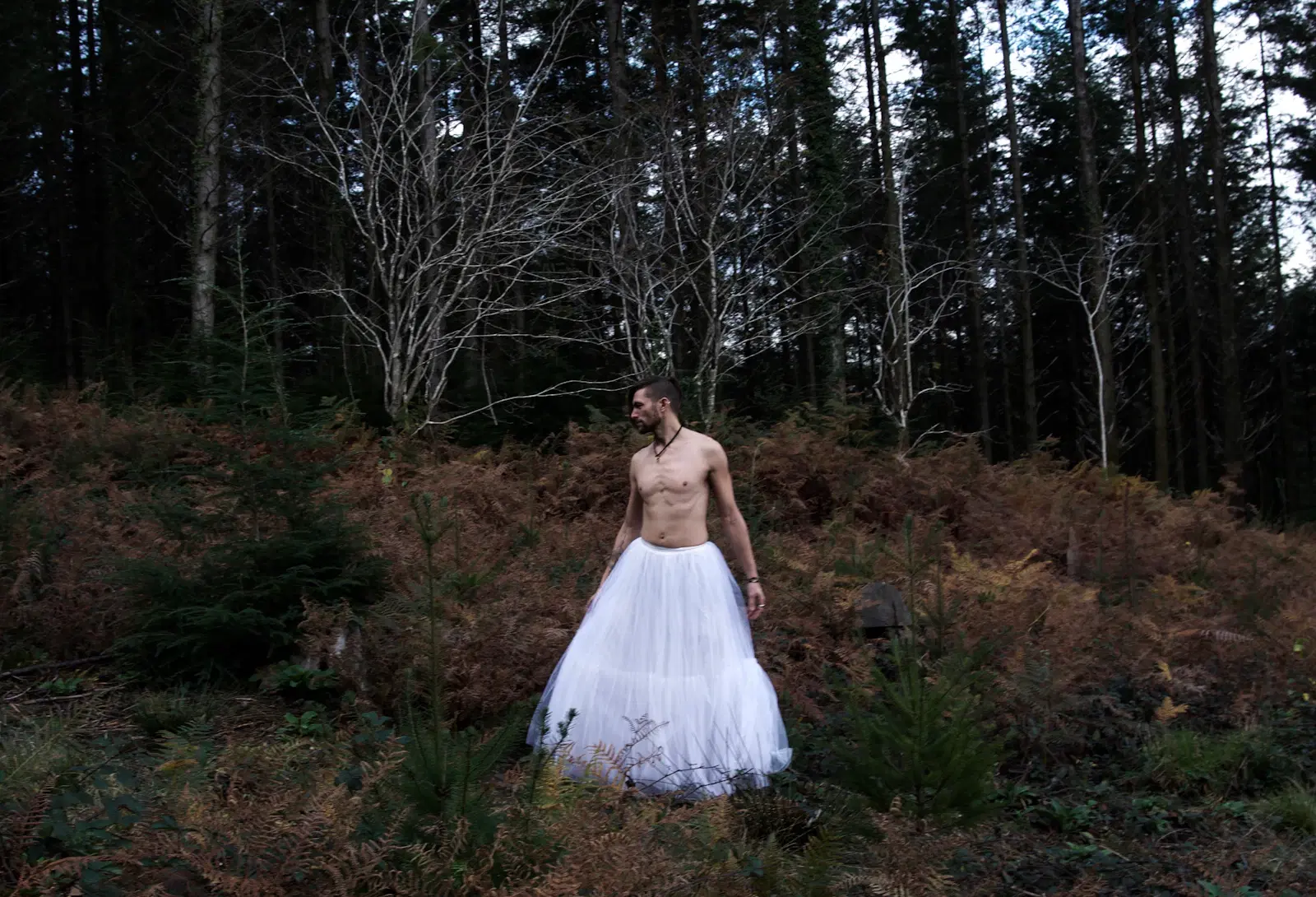 Shirtless man wearing a long white tulle skirt, standing among ferns in a forest