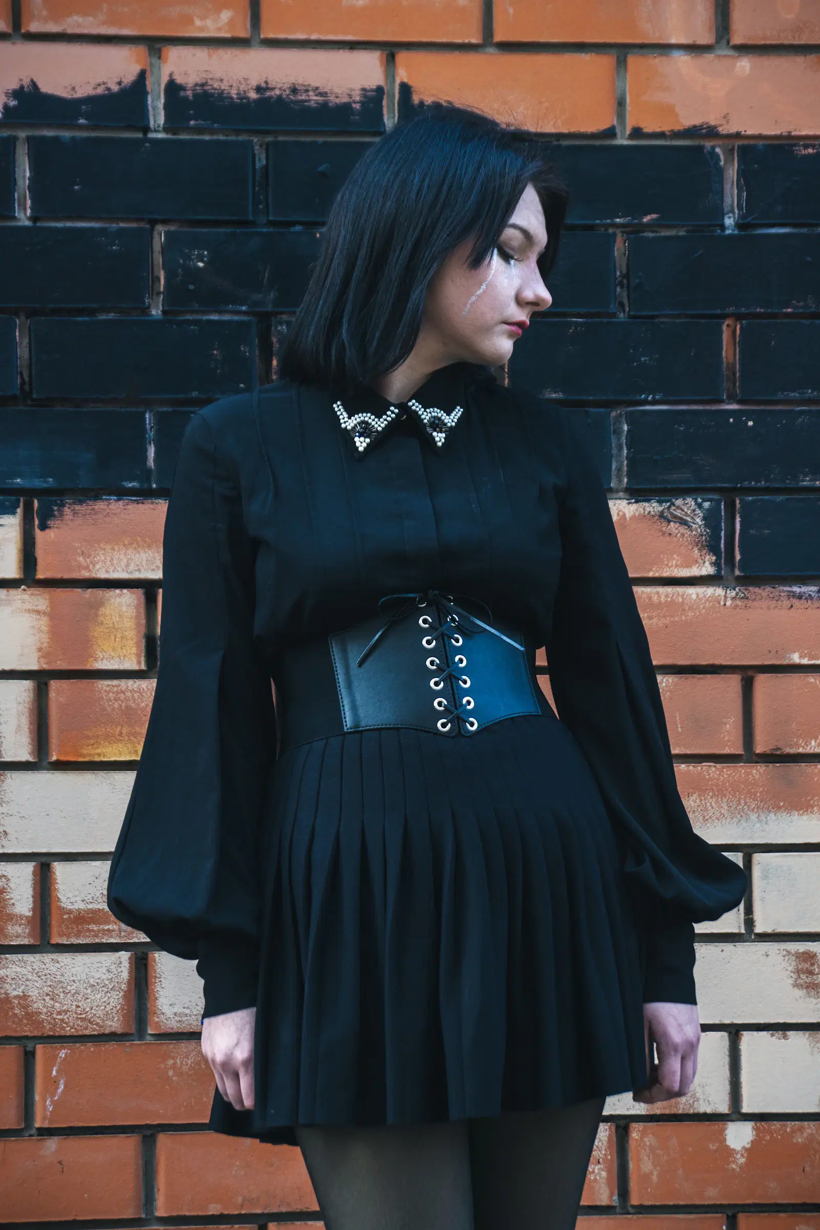 All-black goth outfit with a pleated skirt, wide lace-up belt, black tights, and a blouse with a beaded collar