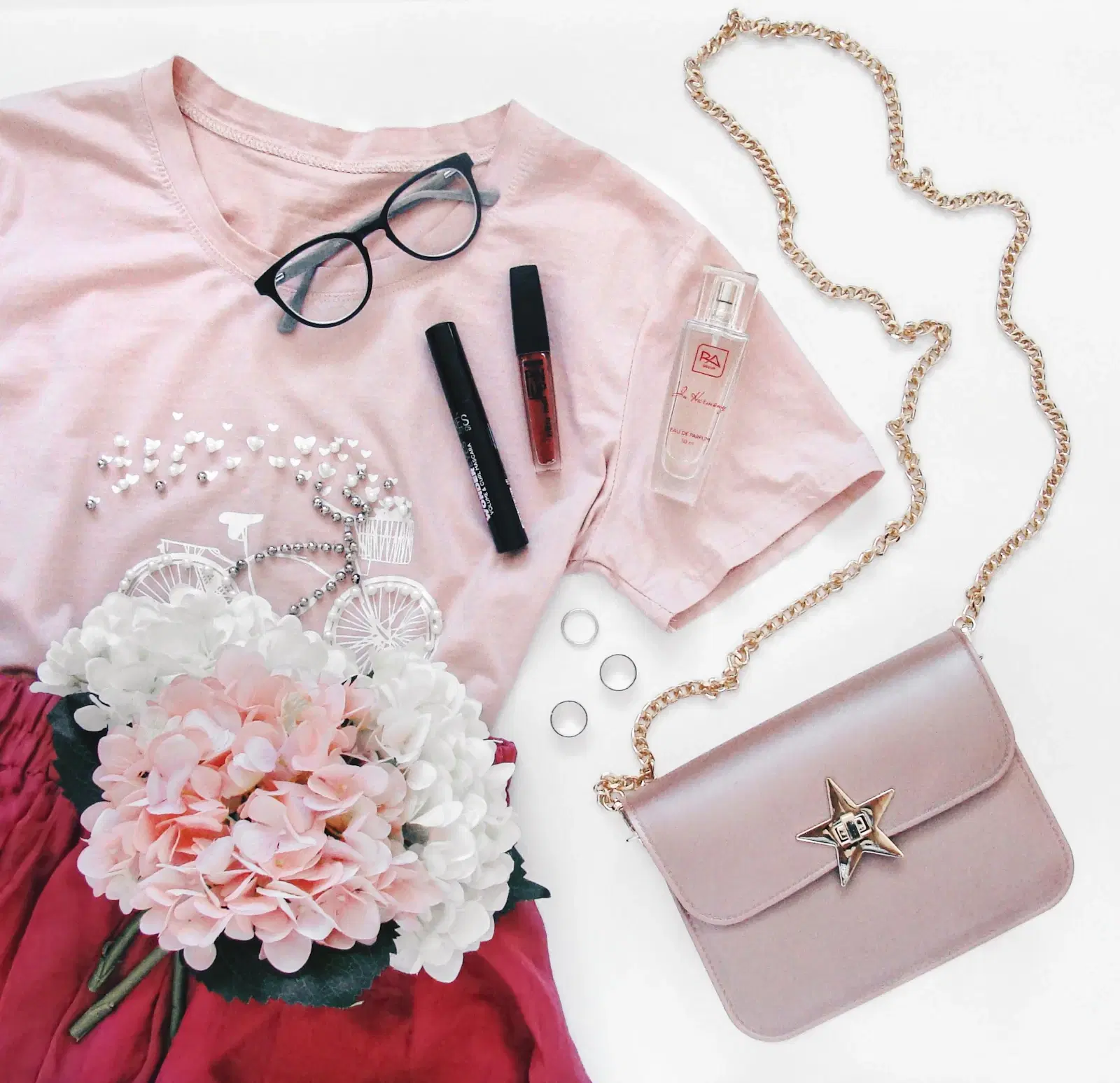 Flat lay of a pastel pink beaded tee, pink chain crossbody bag, glasses, makeup, and hydrangeas