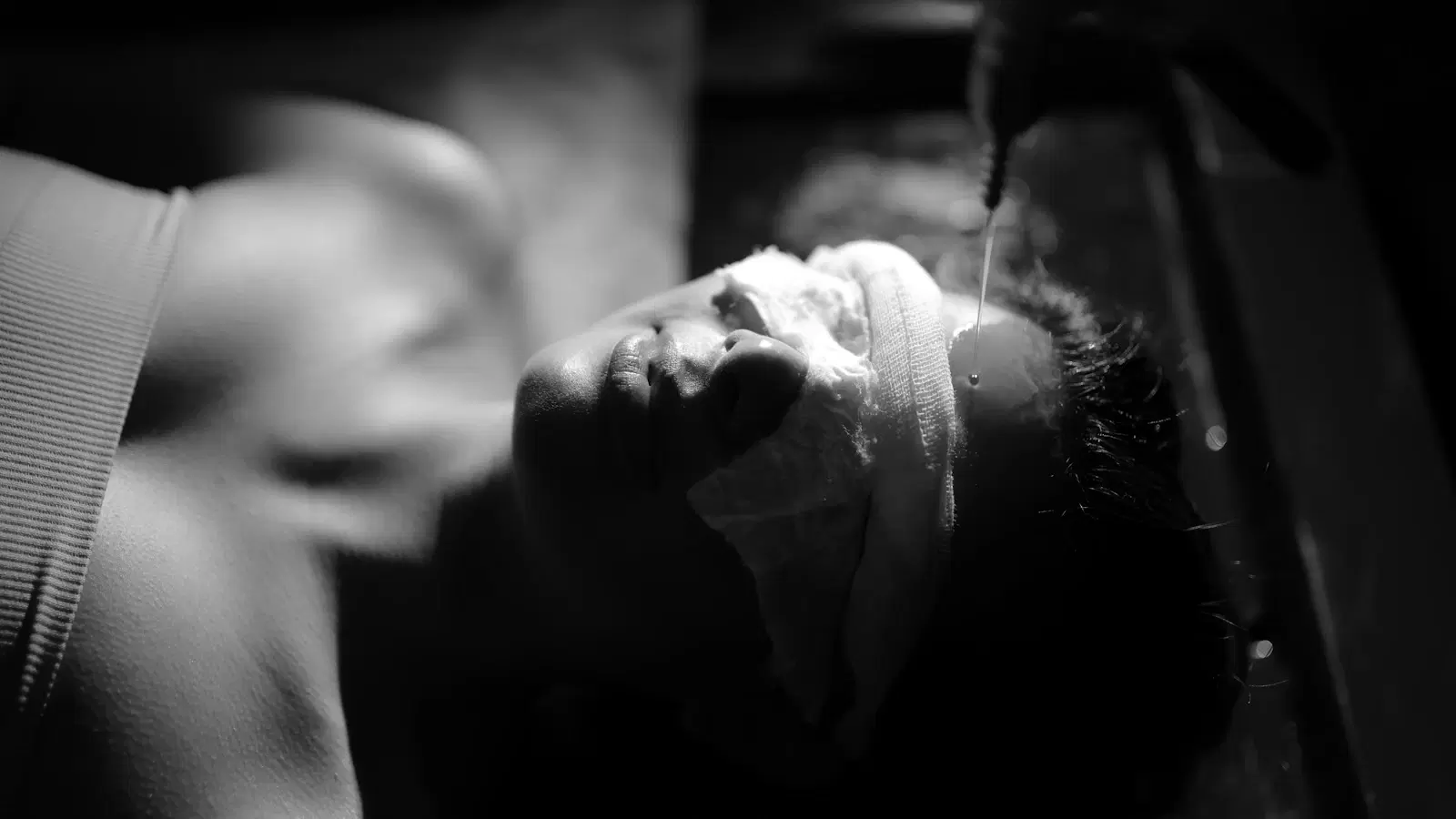 Man seated in a threading salon chair while a technician works on his brows — skin anchored by the client's fingers, thread in the technician's mouth for tension
