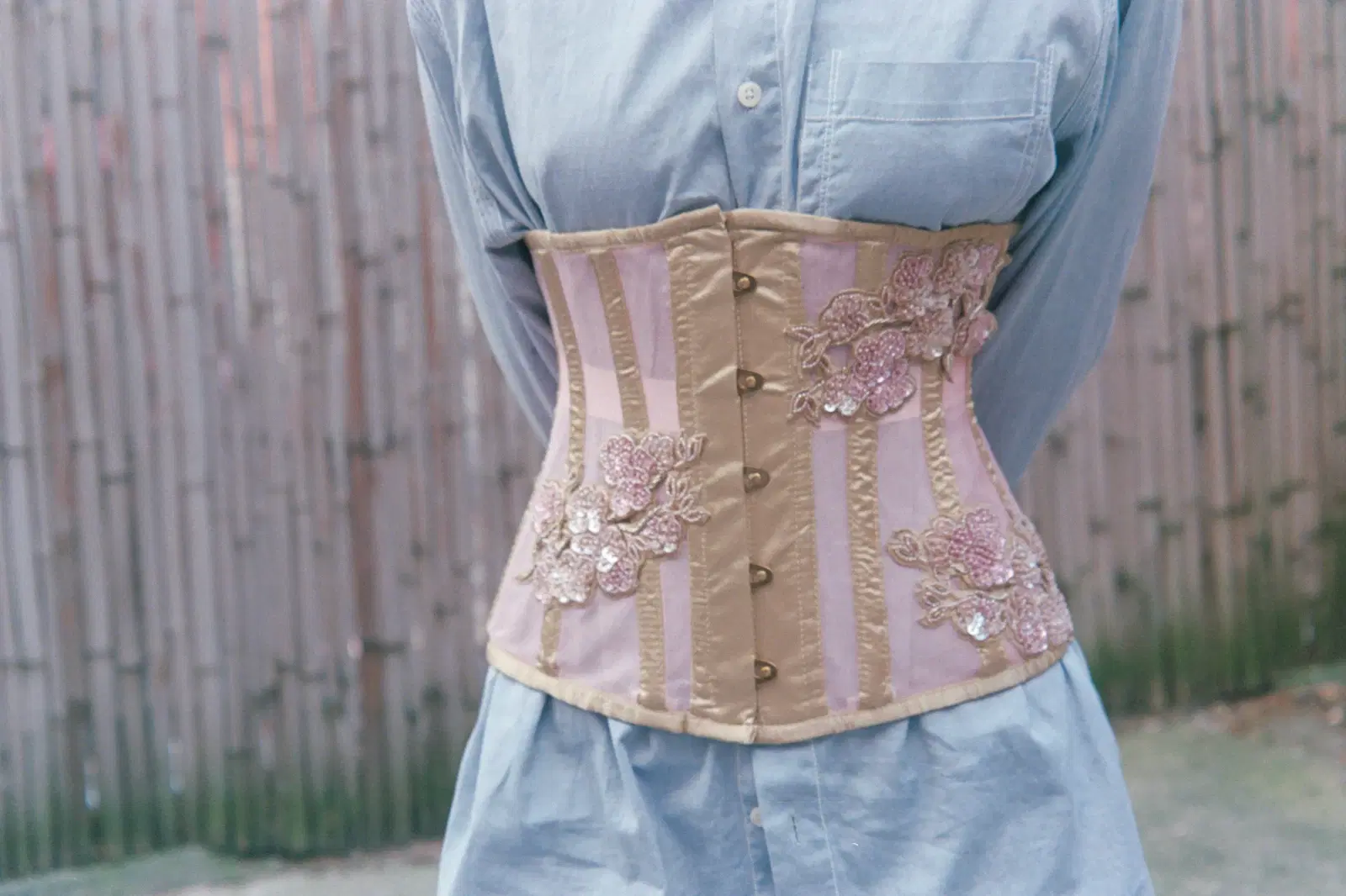 Pink and gold underbust corset with beaded floral appliqués worn over a light blue button-down shirt to cinch the waist