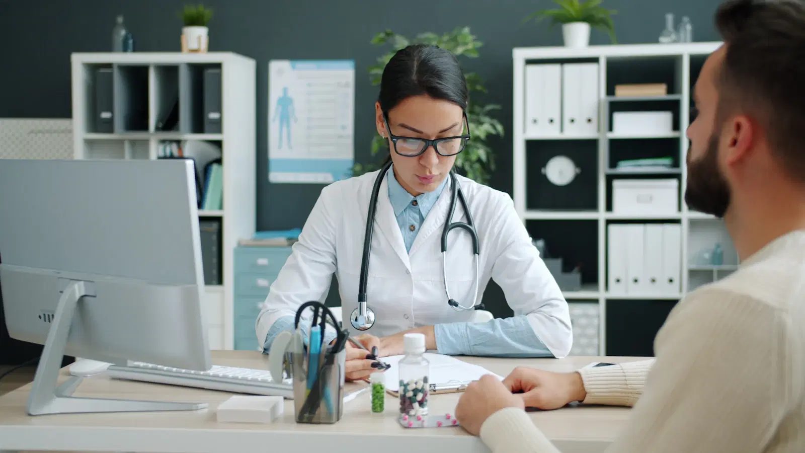 Doctor in a white coat taking notes while consulting with a male patient at her desk
