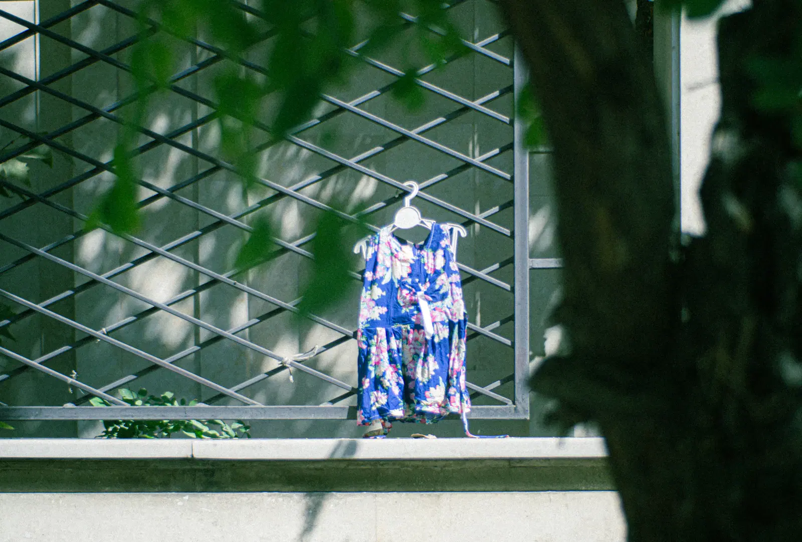 A sleeveless blue floral dress on a hanger, hung from a metal window grille outdoors