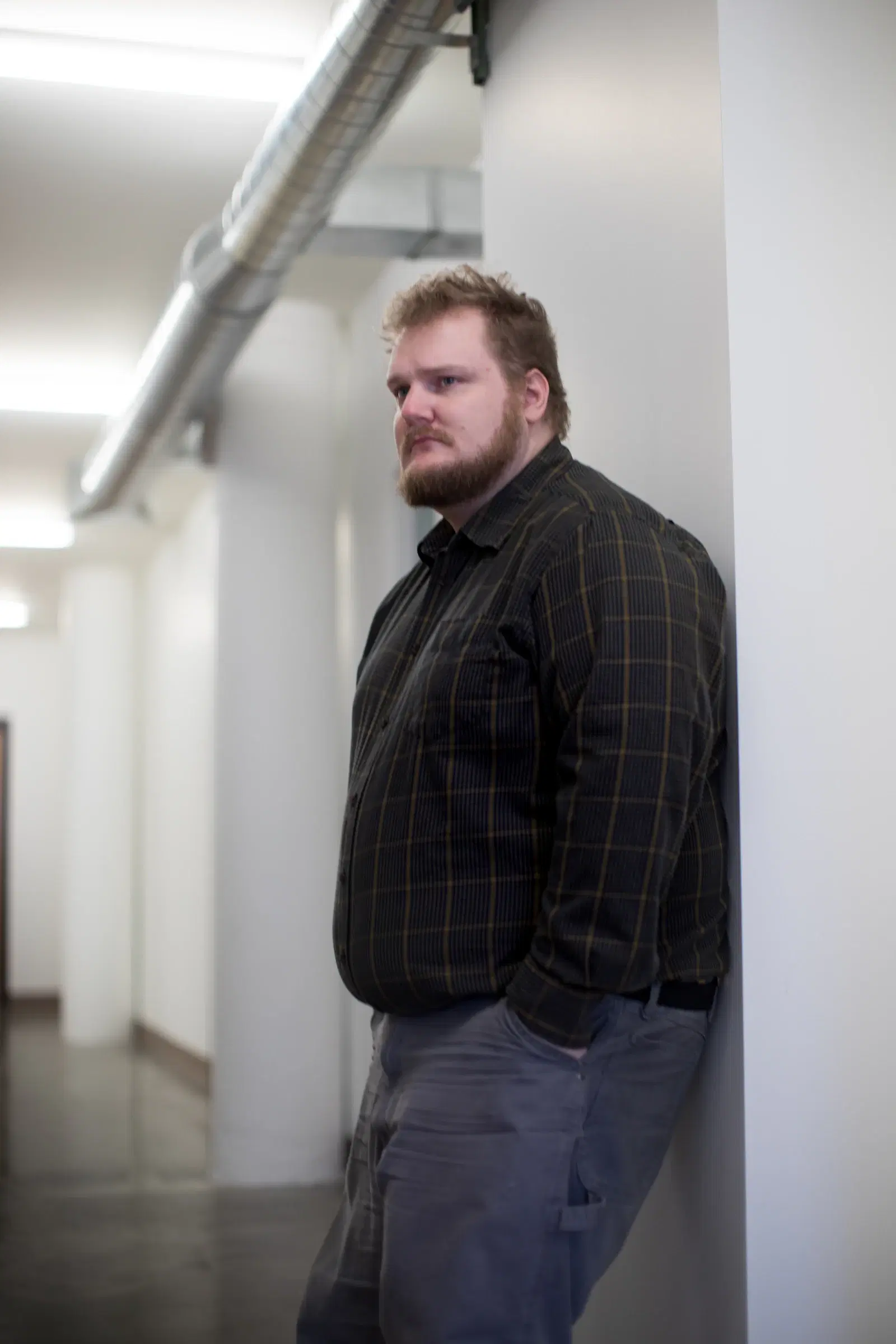 A plus-size man in a dark plaid shirt leaning against a white hallway wall