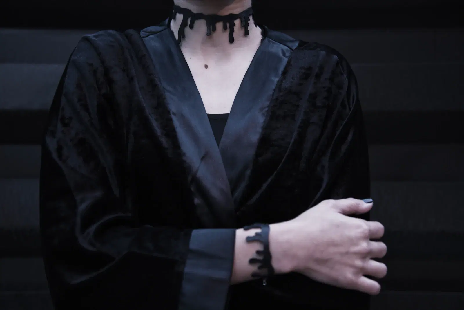 A person in a black velvet robe wearing a black drip-style choker and a matching cuff bracelet