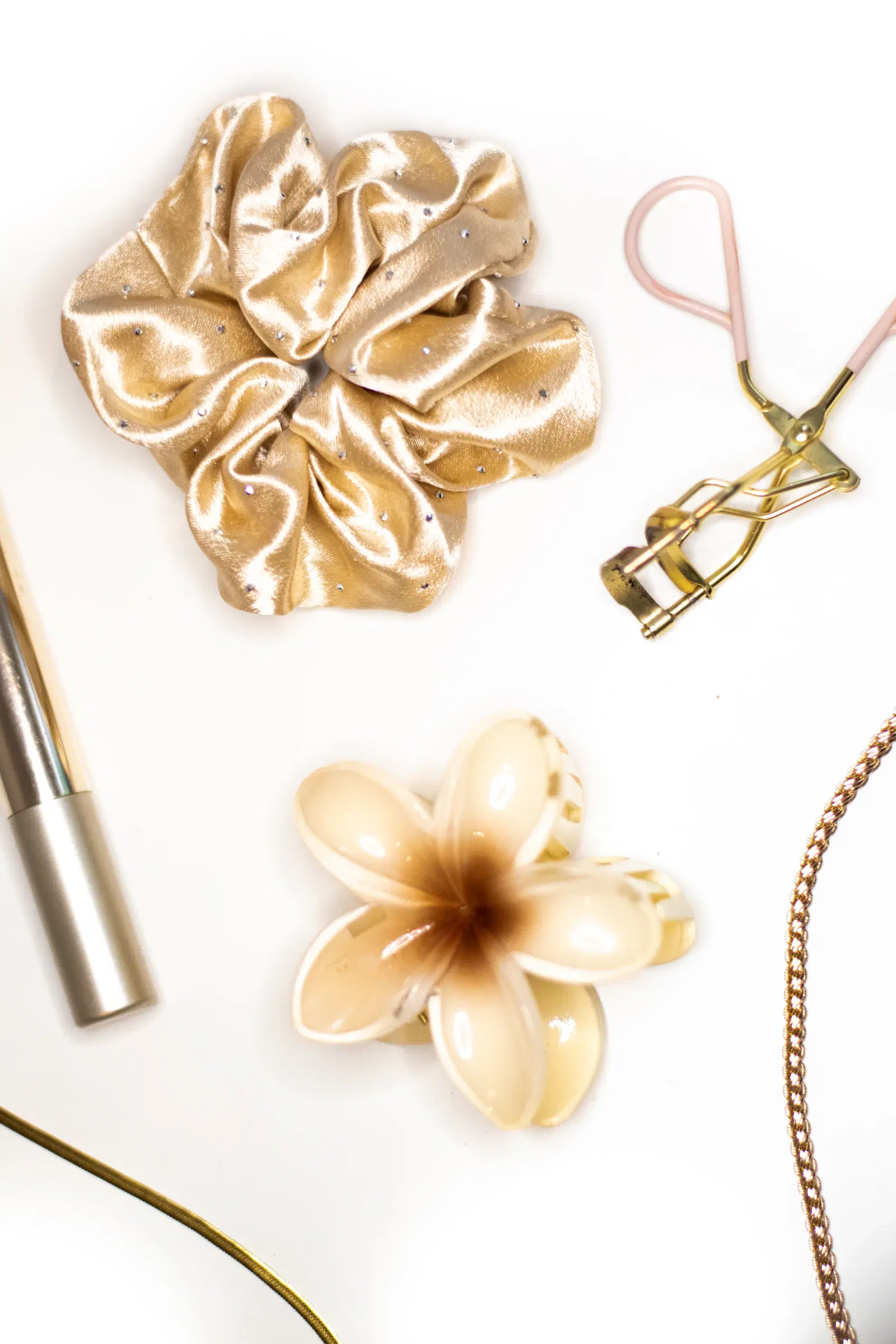 A gold satin scrunchie, a flower claw clip, an eyelash curler, and a gold chain on a white surface
