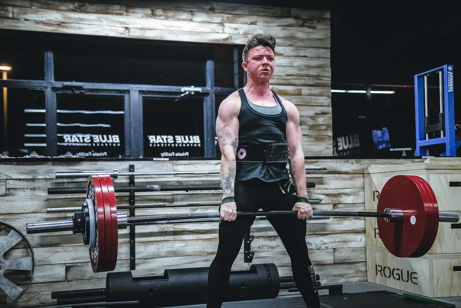 Lifter in a training belt standing tall at the top of a barbell deadlift, a lift that trains the back of the thigh