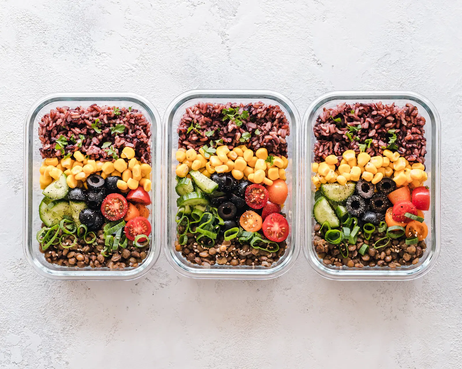 Three meal prep containers of red rice, lentils, corn, and vegetables, the kind of batch cooking that supports thigh growth